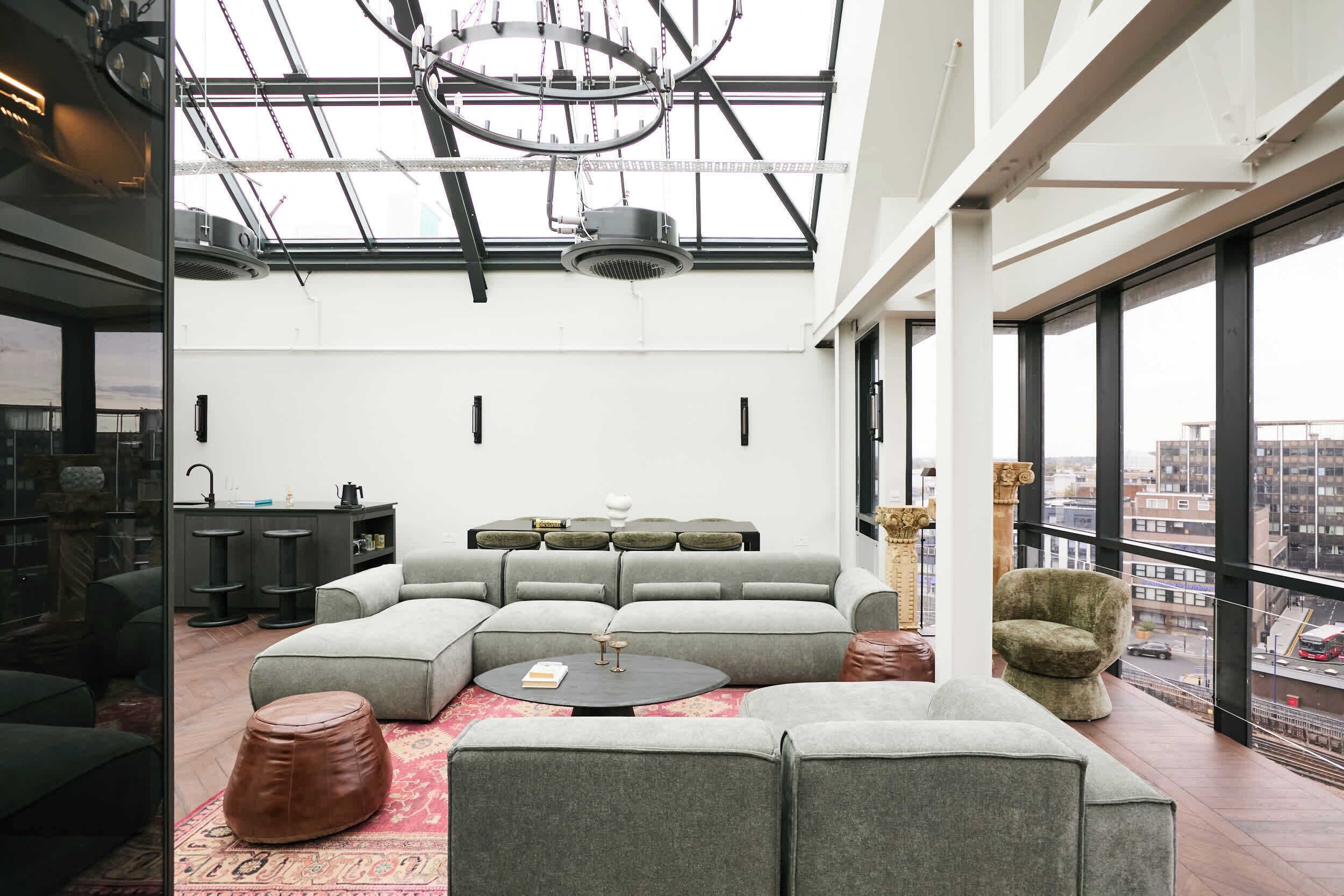 Een moderne woonkamer met een hoekbank, poefs, een salontafel en een stadszicht door grote ramen.