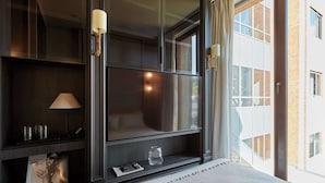 Ein modernes Hotelzimmer mit großem Fenster, einem Bett mit weißer Bettdecke, einem dunklen Holzregal mit Lampe und einem kleinen Tisch mit Glas und Zeitschrift.