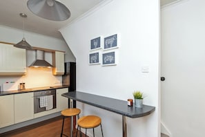 A modern kitchen with white cabinets, a black countertop, and a hanging light fixture. There are two stools and a small potted plant on the counter. - Toast Stays · River-Twine (Croxdale)