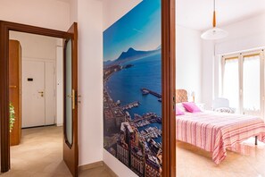 A bedroom with a bed, a nightstand, a lamp, and a view of a coastal cityscape through a large window. - Casa MariaPia al Vomero by Napoliapartments (Naples)