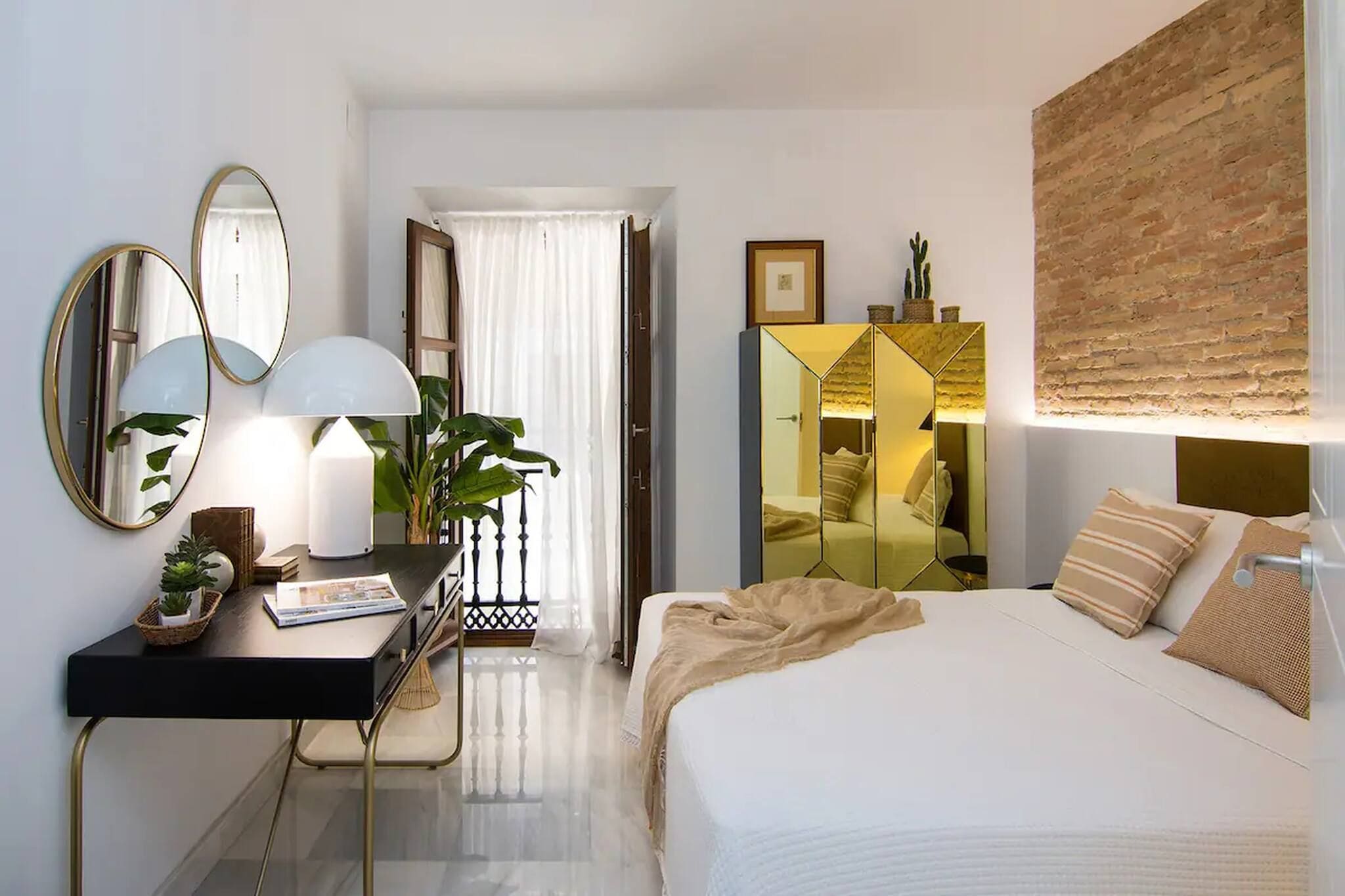 A hotel room with a bed, a desk with a lamp, a mirror, a wardrobe, and a plant.