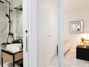 A modern bathroom with a glass shower enclosure, a white sink with a round basin, and a small bedside table with a lamp. - Apartment Riad Alhambra 1 (granada)