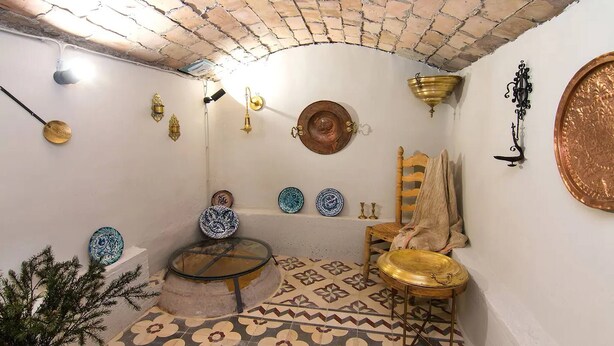 Una stanza con pavimento piastrellato, una sedia in legno, un tavolino e diversi oggetti fissati al muro, tra cui piatti e una lampada.