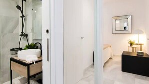 Un bagno moderno con cabina doccia in vetro, lavandino bianco con piano d'appoggio nero e comodino con lampada.