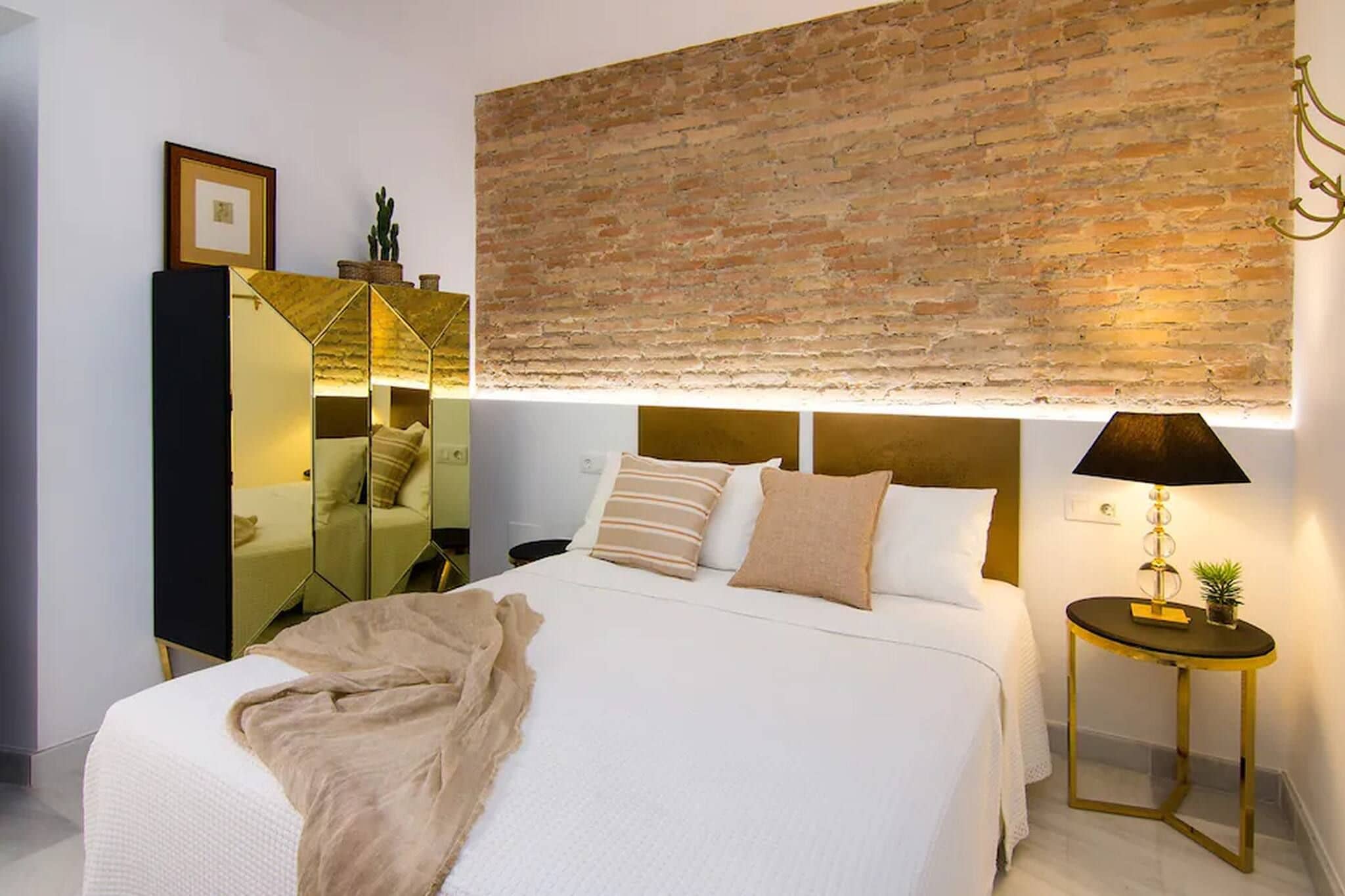 Una habitación de hotel con una cama grande, una mesita de noche con lámpara, un espejo y una pared de ladrillo.