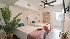 Habitación de hotel con dos camas, un ventilador de techo, un televisor montado en la pared y una planta grande en maceta.