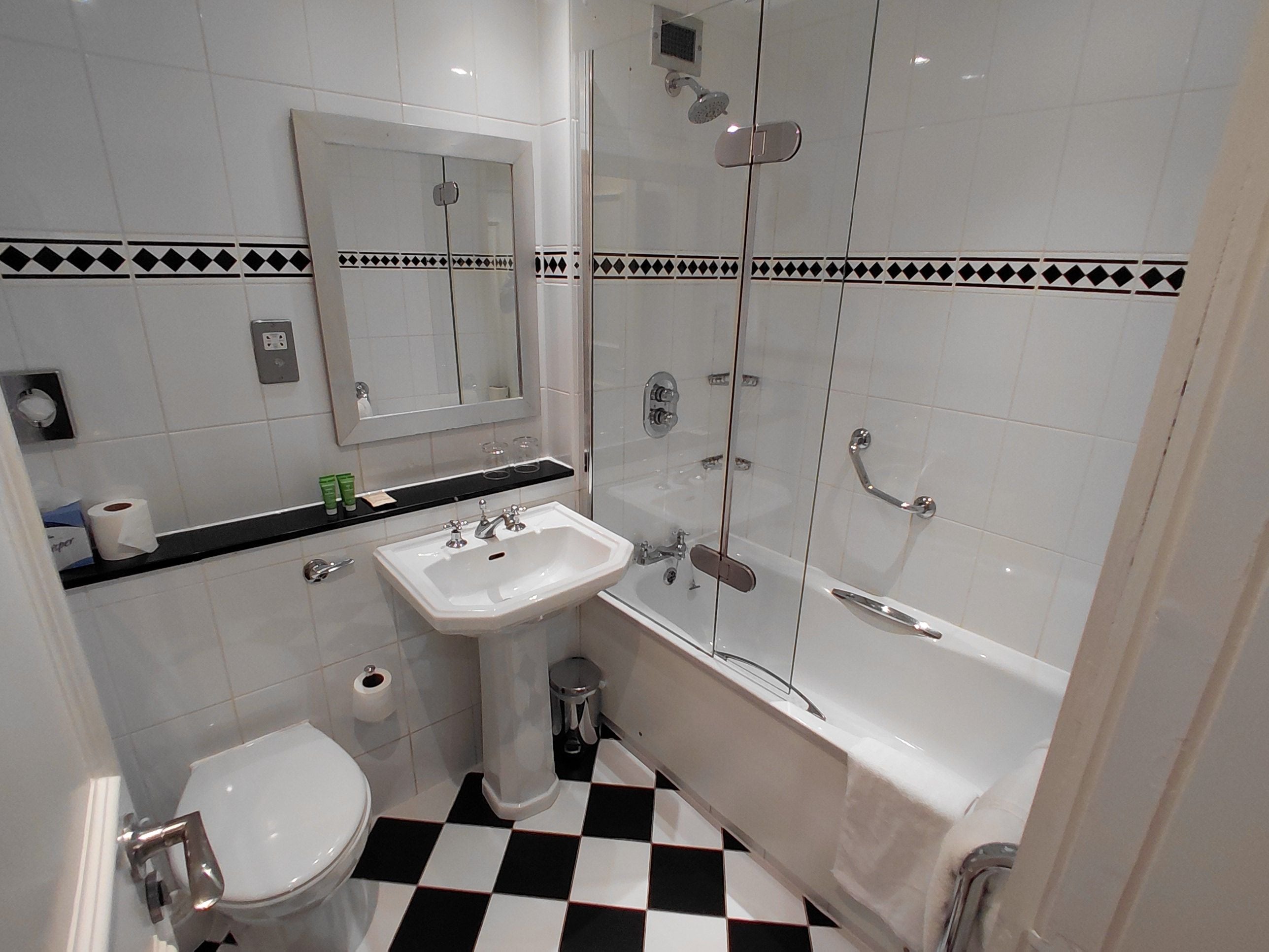 A bathroom with a white sink, toilet, and bathtub. There is a mirror above the sink and a shower with a glass door. The floor has a black and white checkered pattern.