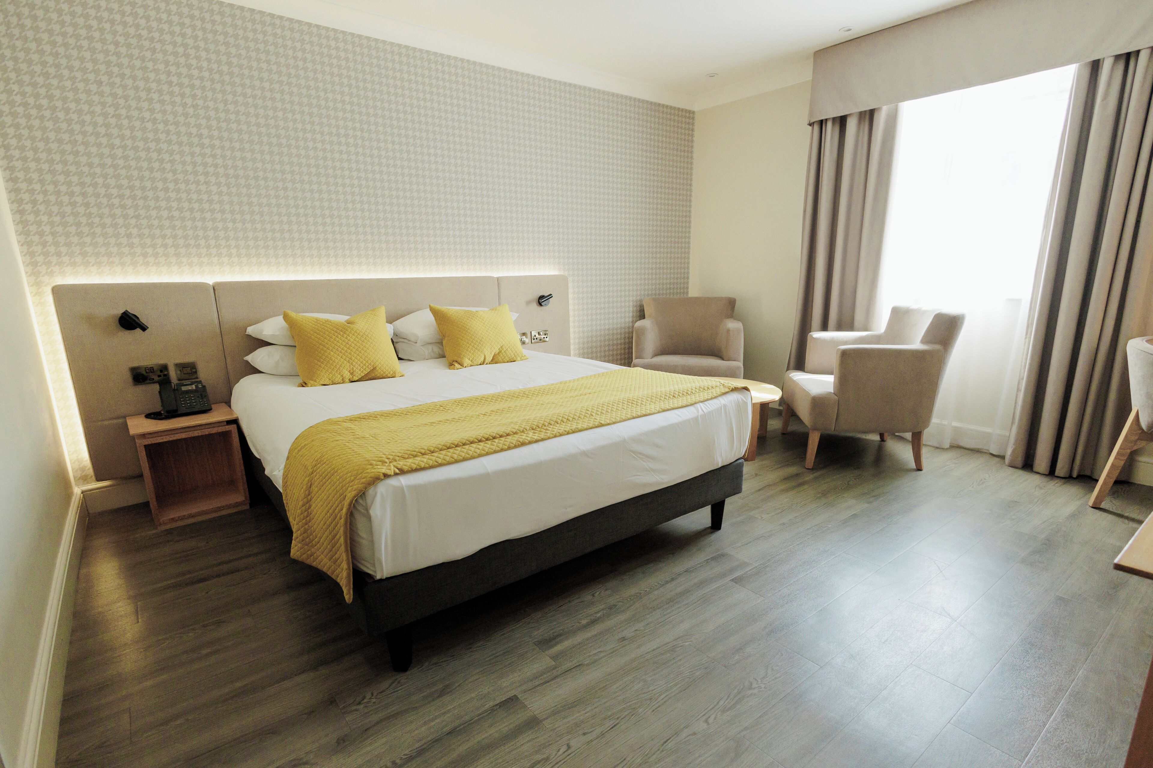 Habitación de hotel con cama, dos sillas, mesita de noche y un ventanal con cortinas.