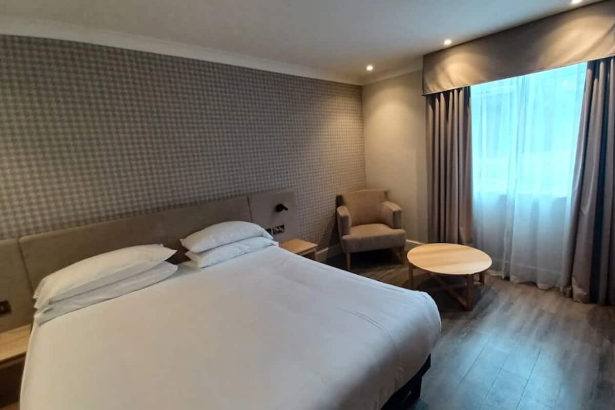 A hotel room with a large bed, a chair, a small table, and a window with curtains.