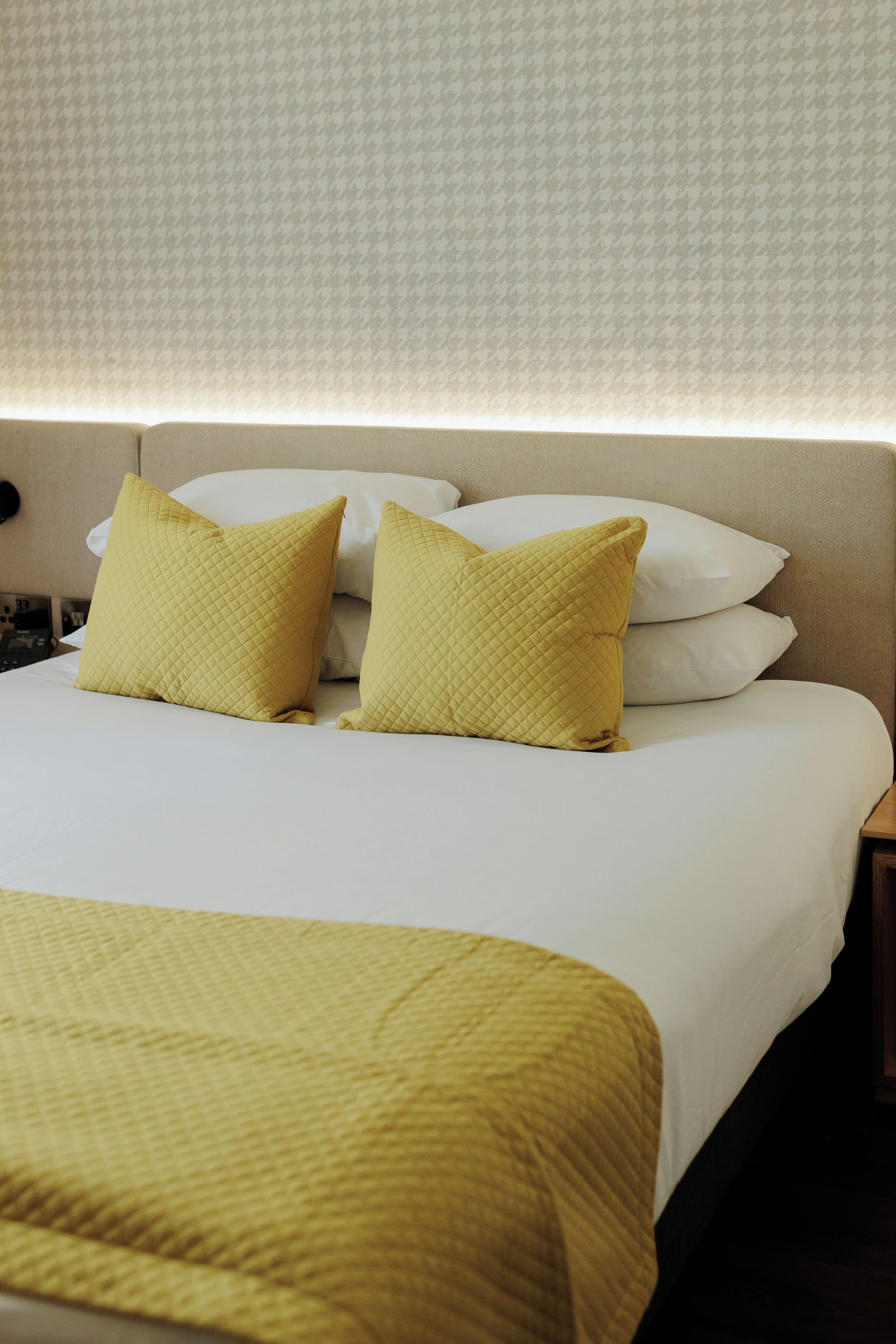 A neatly made bed with white linens and two yellow pillows, a bedside table with a lamp, and a patterned wall in the background.