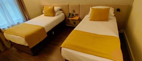 A hotel room with two beds, each with a white mattress and a yellow blanket. There is a nightstand with a phone and a lamp between the beds.