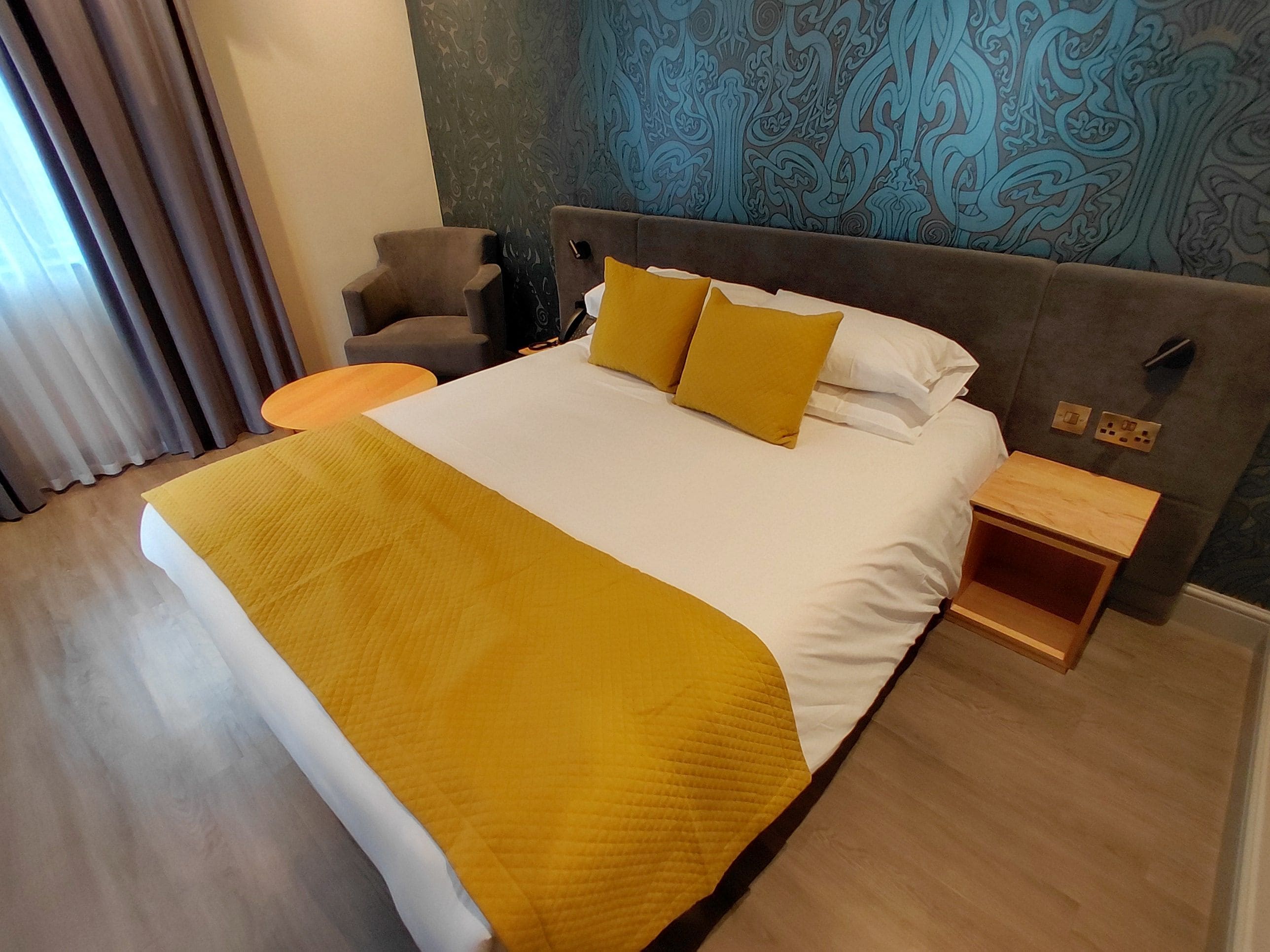 A hotel room with a large bed, a bedside table, a chair, and a patterned wall.