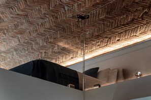 Une pièce avec un plafond orné d’un motif en chevrons, un mur miroir reflétant un lit avec des oreillers, et un luminaire suspendu.