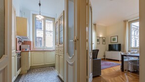 Eine Küche mit Herd, Schränken und einem Fenster. Es gibt eine Essecke mit Tisch und Stühlen, und im Hintergrund steht ein Fernseher.