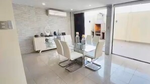 A modern dining area with a glass table and white chairs, a white cabinet with decorative items, and a view through a glass door to an outdoor area. - Stylish Home via Samborondon - No Pets Allowed (Guayaquil)