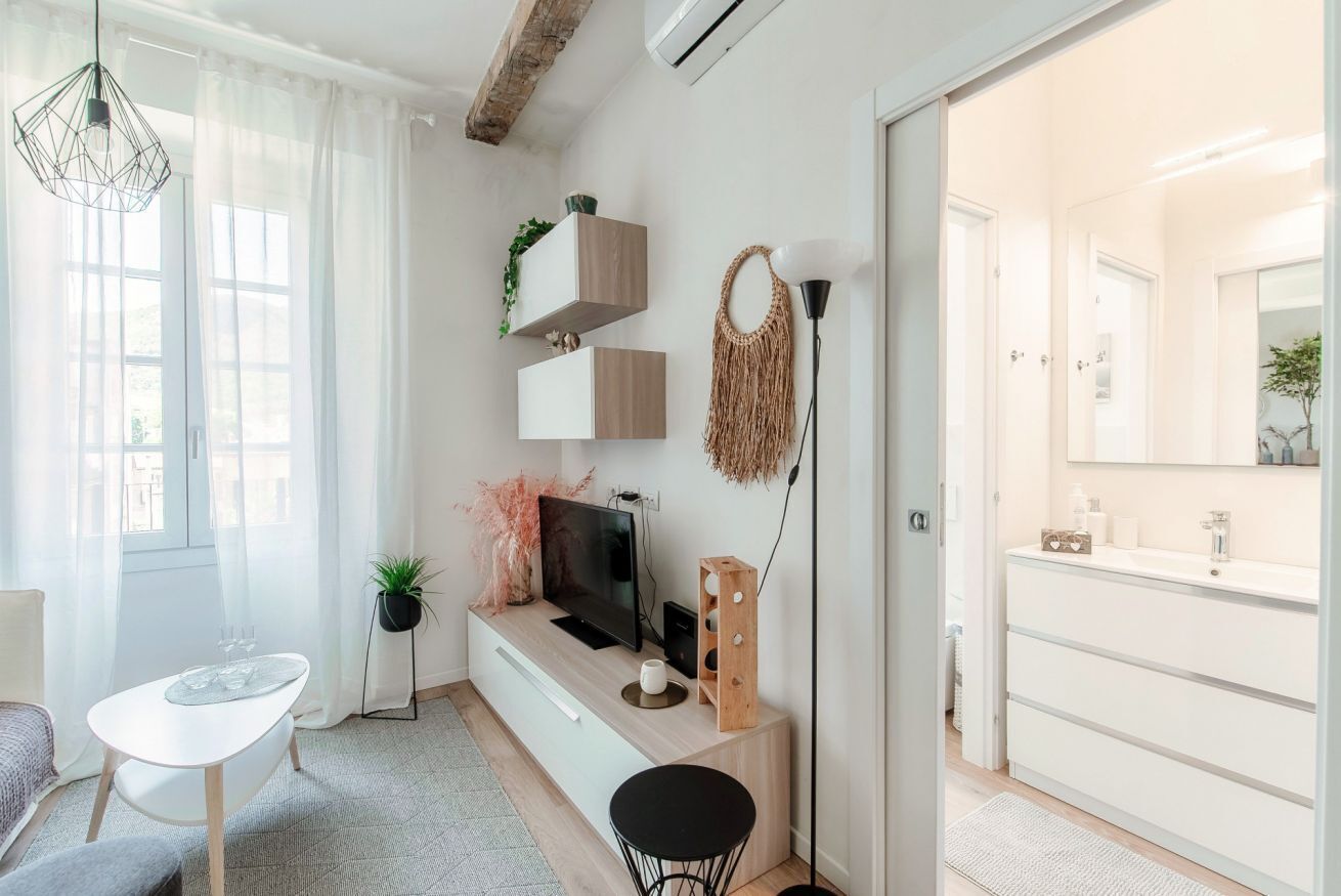 Un salon moderne avec un téléviseur, une petite table à manger et un canapé. Il y a une grande fenêtre avec des rideaux légers, un luminaire suspendu et une plante sur le rebord de la fenêtre.