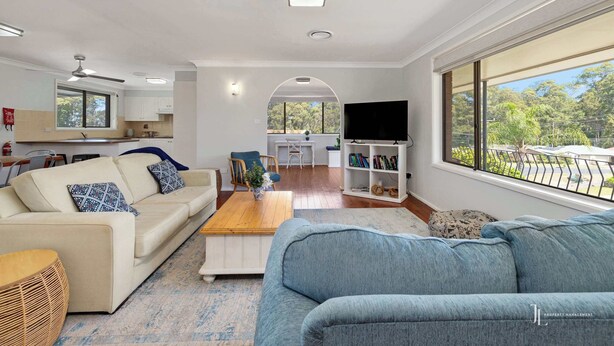 A spacious living area with a sofa, coffee table, and TV stand. There is a kitchen in the background with white cabinets and a window.