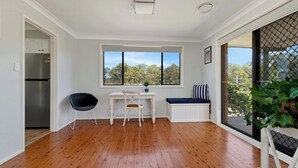 A room with a wooden floor, a white desk with a chair, a bench with a striped pillow, and a view of trees through a window.