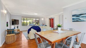 A modern living room with a wooden dining table, grey chairs, a TV, and a large window with a view of houses.