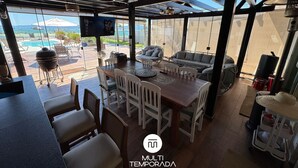Un área de comedor al aire libre con una mesa y sillas de madera, un televisor montado en la pared y vistas a una piscina y follaje.