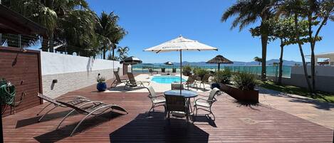 A wooden deck with lounge chairs and a pool, surrounded by palm trees and a clear view of the ocean.