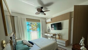 A modern bedroom with a bed, bedside table, TV, and a view of the outdoors. - Dream House Frente Mar - Ponta das Canas (Florianópolis)