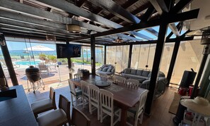 An outdoor dining area with a wooden table and chairs, a TV mounted on the wall, and a view of a pool and greenery. - Dream House Frente Mar - Ponta das Canas (Florianópolis)