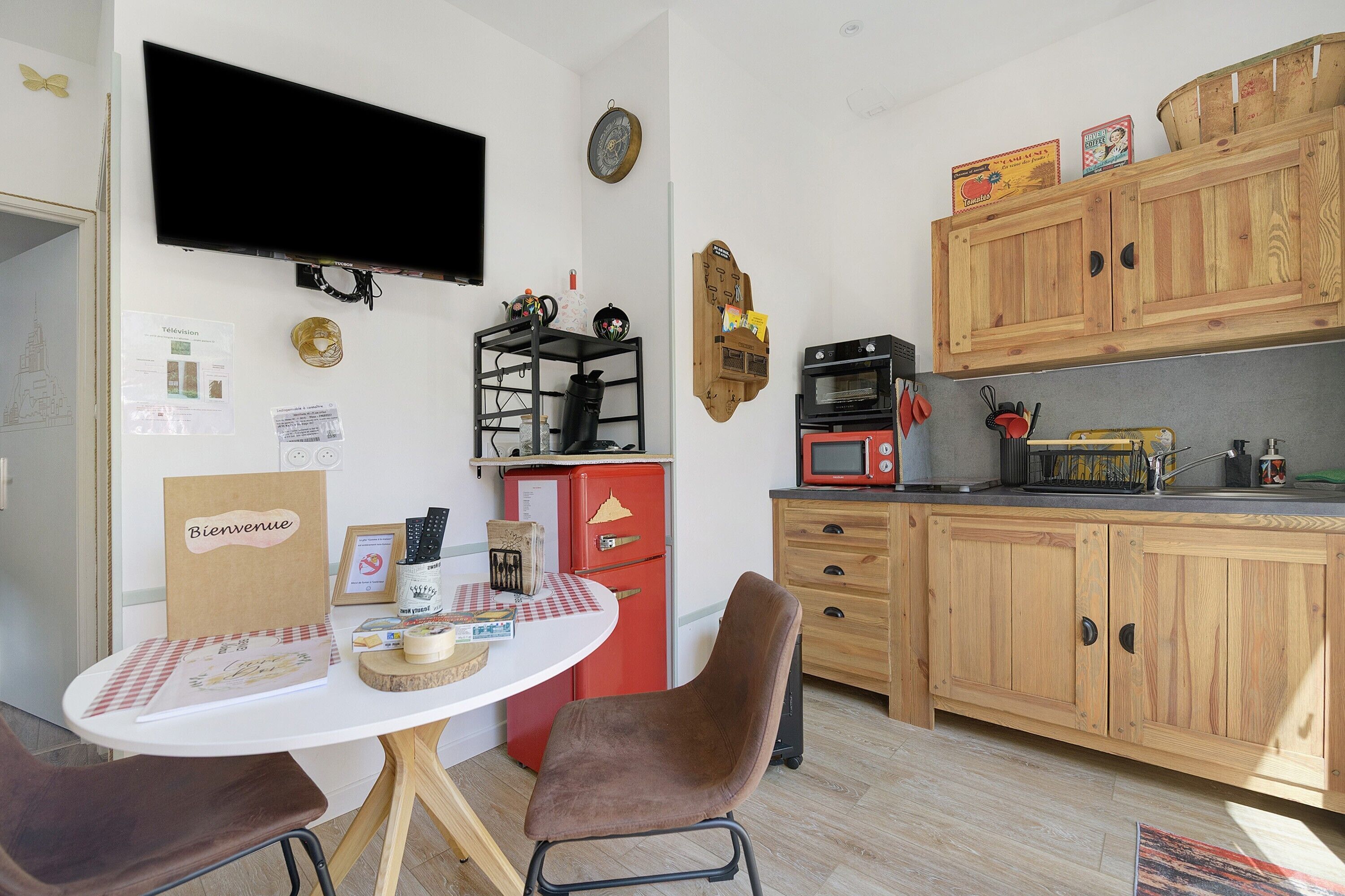 A compact kitchen with a dining table, chairs, a red refrigerator, a microwave, wooden cabinets, and a television mounted on the wall.