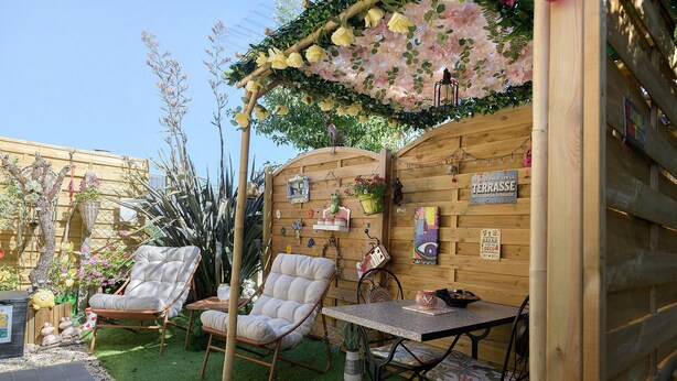 Eine hölzerne Pergola mit hängenden Blumen, ein Holzzaun, ein Tisch mit Stühlen und ein Schild, das eine Terrasse kennzeichnet.