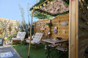 A wooden pergola with hanging flowers, a wooden fence, a table with chairs, and a sign indicating a terrace. - Holiday Home 'Comme À La Maison' with Private Terrace, Garden & Wi-Fi (ducey)