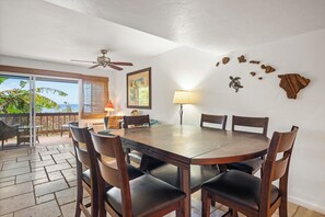 Un coin repas avec une table ronde, six chaises, un ventilateur de plafond, une lampe et une vue sur l’océan, visible à travers une porte-fenêtre coulissante.