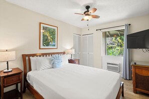 Une chambre à coucher comprenant un lit, un ventilateur de plafond, une table de chevet avec une lampe, un téléviseur et une fenêtre avec des rideaux.