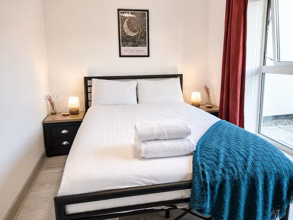 A neatly made bed with white linens and two folded towels, a framed picture above the bed, and a window with red curtains to the right.