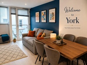 A modern living room with a sofa, dining table, and chairs. There are framed artworks on the wall and a window with blinds. - York Centre Flat - balcony, lift & 2 baths (York)