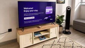 A television displaying a welcome message with a check-out date selection option, a Roku logo, and a wooden entertainment unit with books and a plant. - York Centre Flat - balcony, lift & 2 baths (York)