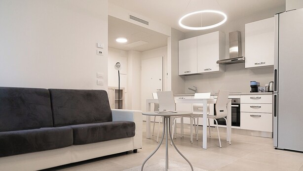 Una sala moderna con un sofá gris oscuro, una mesa de comedor blanca con sillas y una cocina con armarios blancos y un refrigerador.