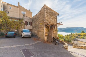 A narrow street with parked cars, a stone building with a tree, and a view of a lake and hills. - L'amourié, ground floor, 4-person apartment with lake view (Sainte-Croix du Verdon)