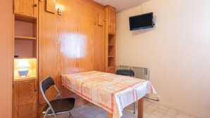 Ein kleines Zimmer mit einem Holzkasten, einem Tisch mit Tischdecke, einem Stuhl, einem Heizkörper und einem an der Wand montierten Fernseher.