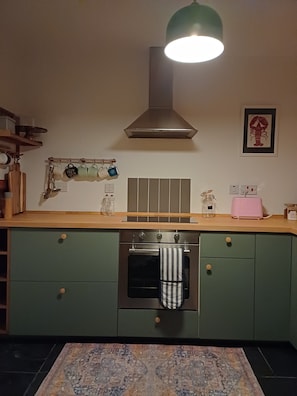 A kitchen with green cabinets, a stainless steel oven, a hanging rack with mugs, and a tiled backsplash. - Cosy Scottish Highlands Acharacle Cottage, Amazing Views, Village Location (Acharacle)