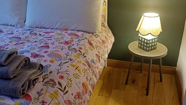 Siististi pedattu sänky, jossa on kukkakuviollinen päiväpeite ja tyynyjä, pieni pyöreä pöytä, jossa on lamppu, ja puulattia.