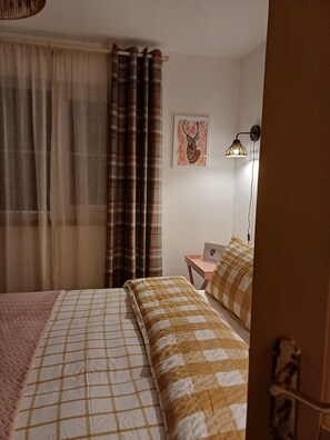 Ein Schlafzimmer mit zwei Betten, einem Nachttisch mit Lampe, einem gerahmten Bild an der Wand und einem Fenster mit Vorhängen.