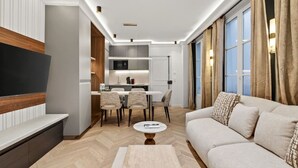 A modern living room with a sofa, a coffee table, and a TV. There is a dining area with chairs and a table. The room has wooden flooring and large windows with curtains. - New suite 6 personnes- Saint Germain Notre-Dame (Paris)