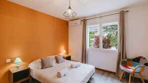 Un dormitorio con cama, lámparas de noche, unidad de aire acondicionado, ventana con cortinas y una silla.