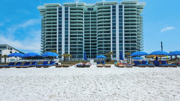 Um hotel moderno com várias varandas, uma área de praia com espreguiçadeiras e guarda-sóis, e um céu azul límpido.