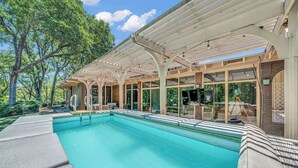 A swimming pool with a covered patio area, surrounded by trees and a clear sky. - Luxe Austin Retreat, Pool & Yard, Sleeps 20 (Austin)