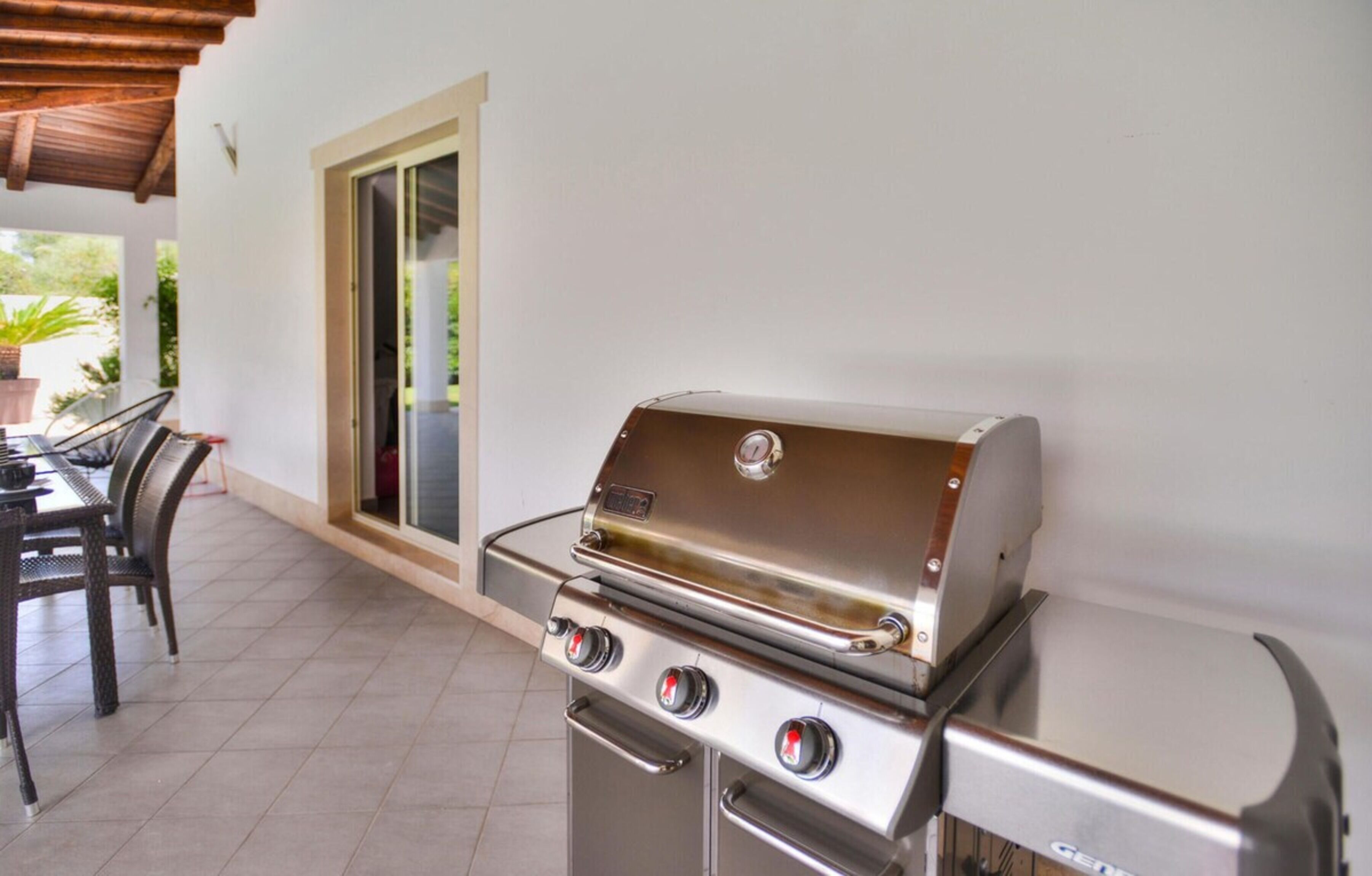 A patio with a stainless steel grill, a dining table with chairs, and a sliding glass door leading to a garden.