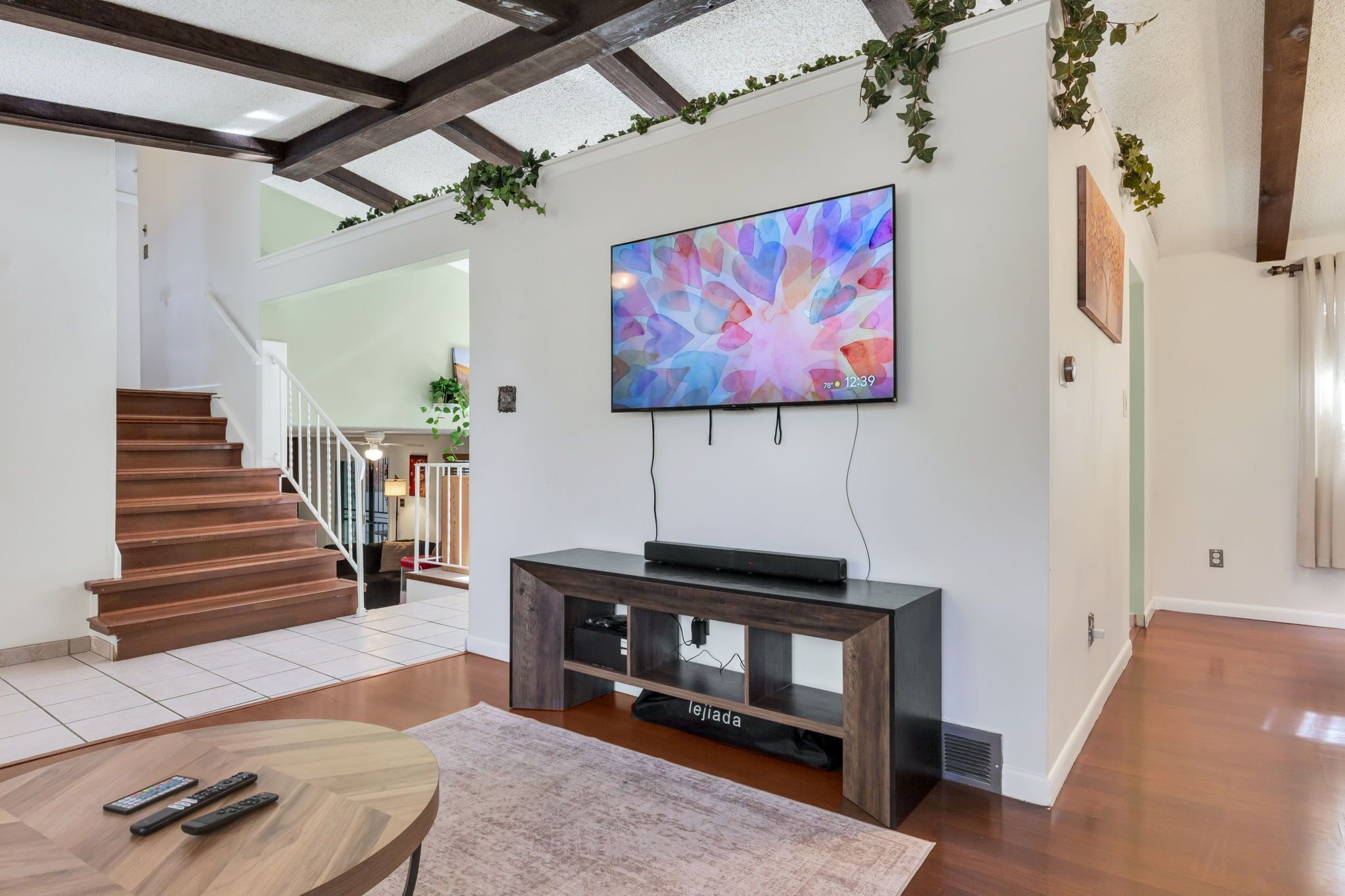 A modern living room with a wooden staircase, a flat-screen TV mounted on the wall, and a console table with a remote control.
