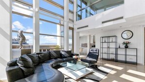 A modern living room with a leather sofa, a glass coffee table, and a black armchair. There is a large window providing natural light and a view of the city. - Welcome to The Penthouse – Beachfront Luxury (Barcelona)