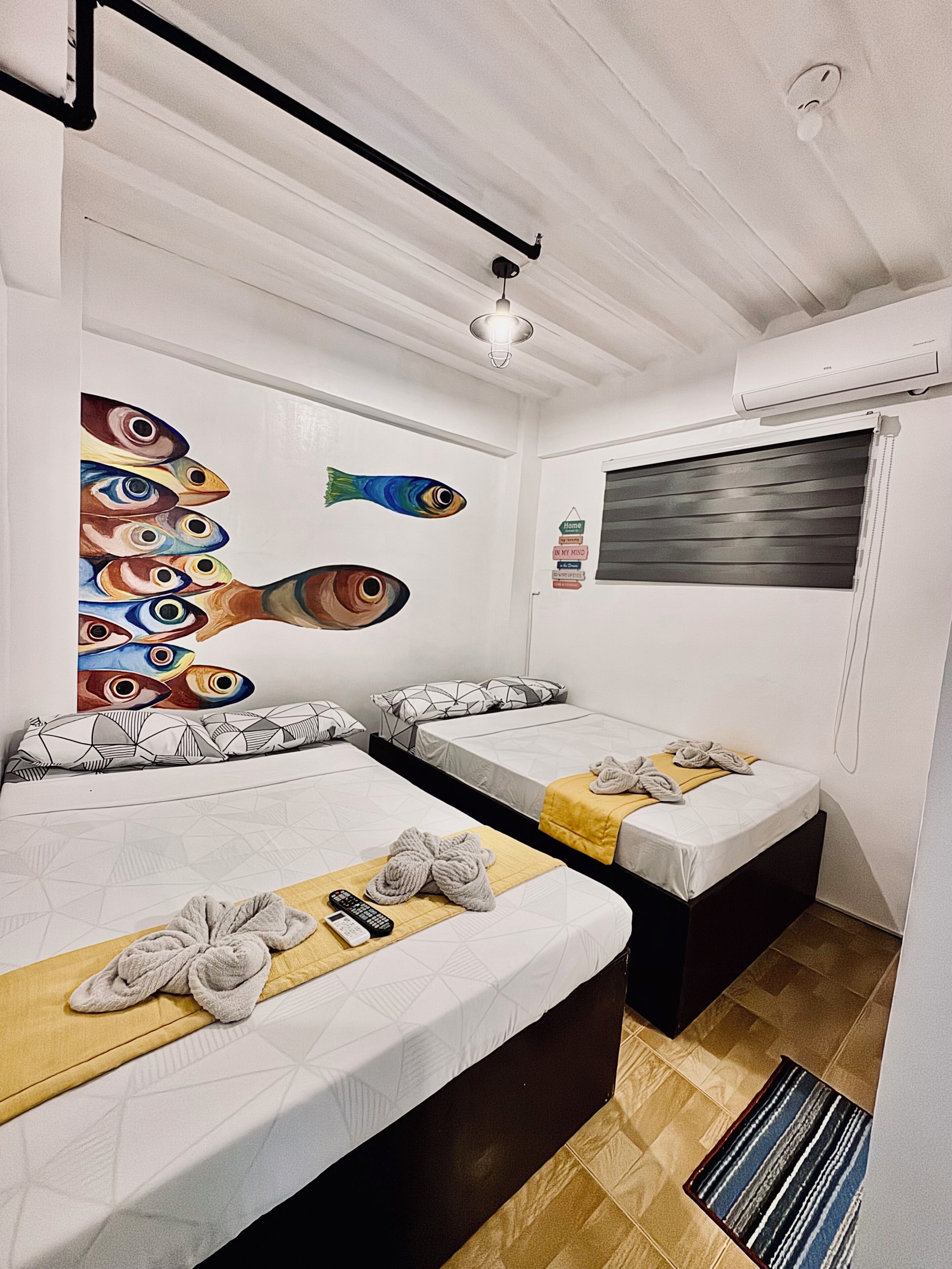 A hotel room with two beds, a ceiling fan, a painting of fish, a window with blinds, and a small rug.