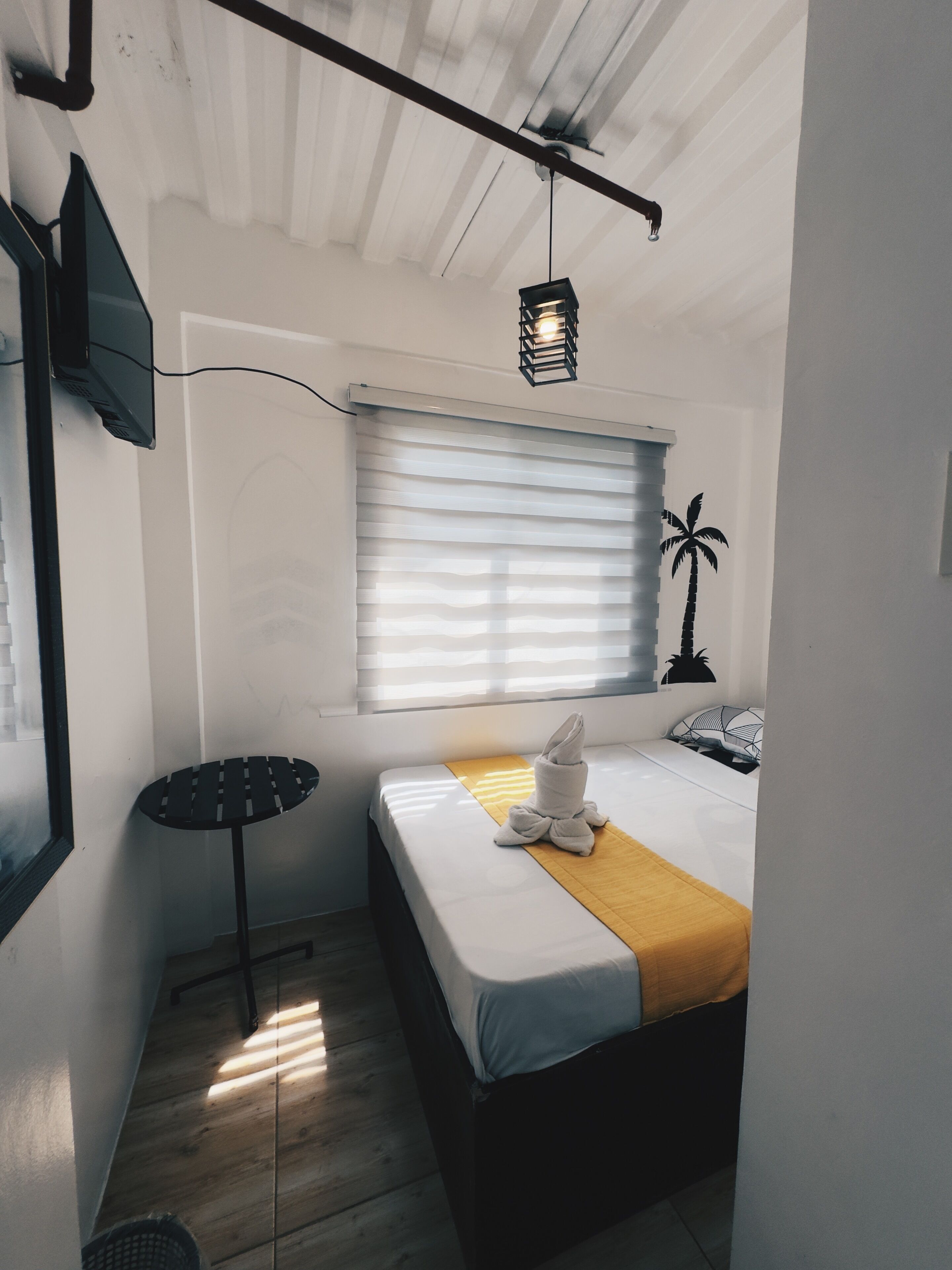 A compact hotel room with a single bed, a small table, a TV mounted on the wall, and a window with blinds.